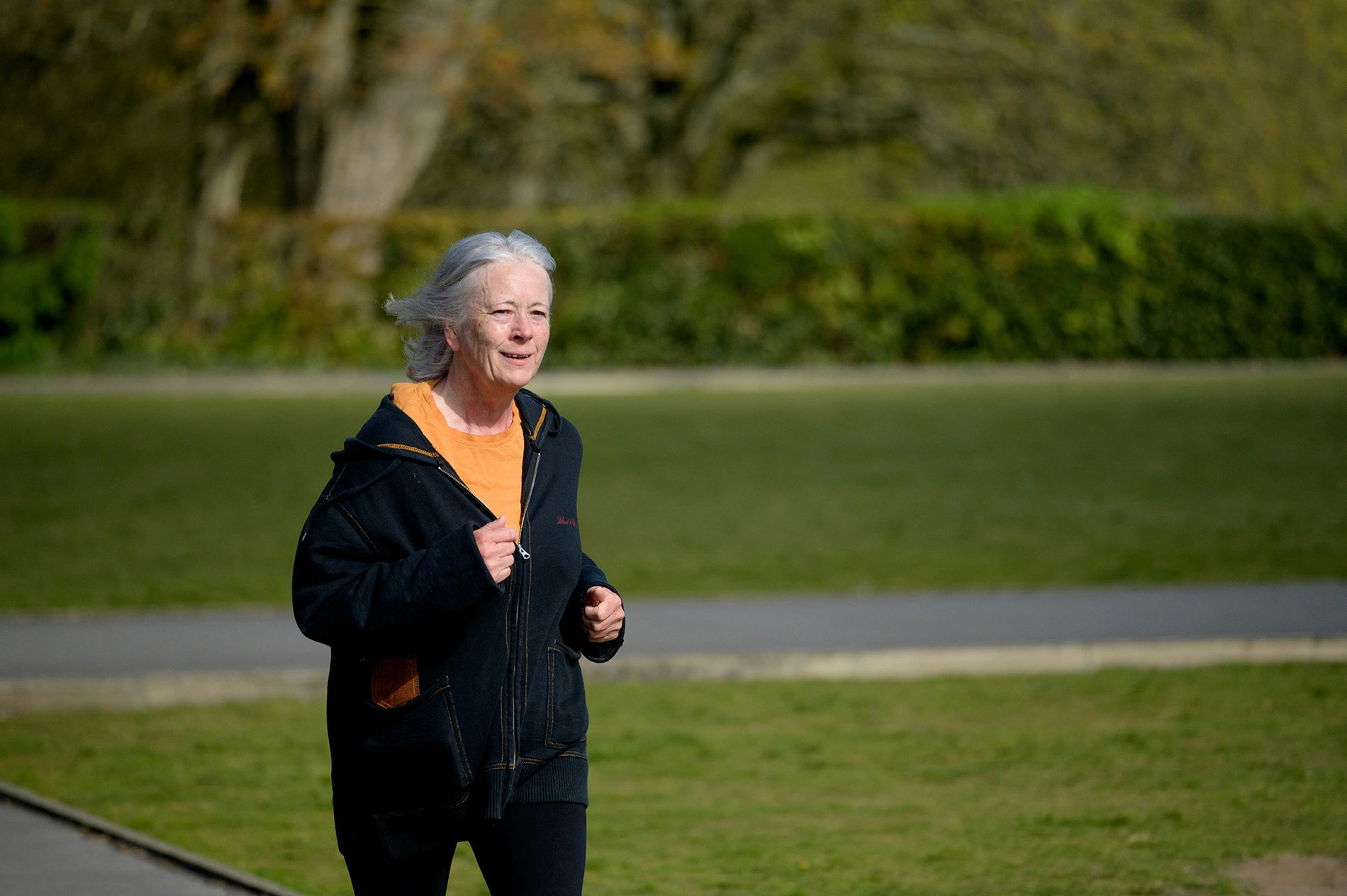 Older lady running at a park in black and orange athletic gear while she's smiling